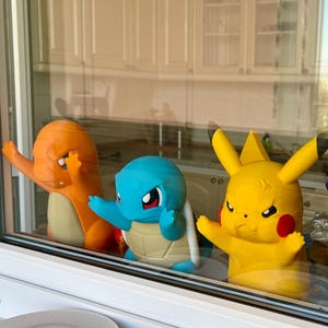 May include: A window display featuring four colourful 3D-printed Pokémon figures: Charmander, Squirtle, Pikachu, and Gengar. The figures are in vibrant orange, blue, yellow, and purple, with detailed features and expressions. The figures are displayed in front of a window.