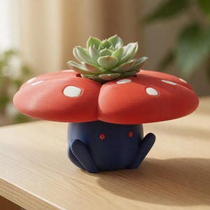 Op de afbeelding: Een grillige paddenstoelvormige bloempot in rood en blauw, met witte stippen op de kap. Een groene vetplant staat bovenop de pot. De pot staat op een houten oppervlak.