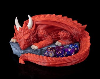 May include: A red dragon figurine resting on a gray stone base, with white horns and claws. Several multi-sided dice in shades of blue and purple are nestled beside the dragon, suggesting a tabletop gaming theme.