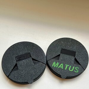 May include: Two black, circular objects with a textured surface. One has the word "MATUS" printed in green. Each object has a rectangular cutout and a triangular section removed, creating a unique shape.