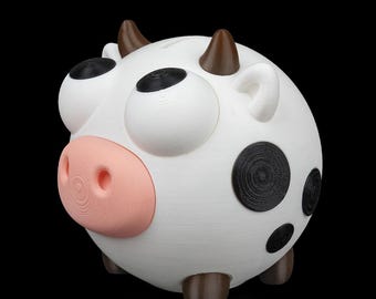 May include: A whimsical, 3D-printed piggy bank shaped like a cow. The white bank features large, black-pupiled eyes, black spots, and a pink nose. Brown horns and feet complete the design. The bank is a fun, decorative item.