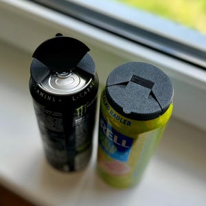 May include: Two aluminum beverage cans, one black and one green, each topped with a black plastic lid. The black can has white text and a green logo. The green can has yellow and blue text.