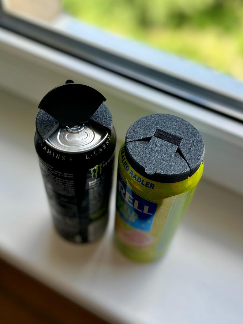 May include: Two aluminum beverage cans, one black and one green, each topped with a black plastic lid. The black can has white text and a green logo. The green can has yellow and blue text.