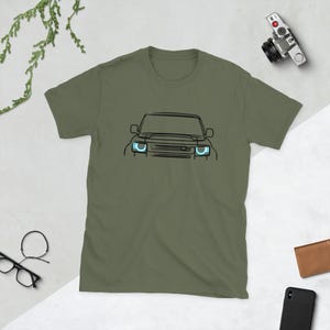 May include: A green short-sleeved t-shirt with a black line drawing of a car's front end. The car has round headlights with a blue outline.