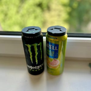 May include: Two aluminum beverage cans, one black with green claw marks and the word "MONSTER", the other yellow with the word "RELL". Both cans have black plastic lids. The background is a blurred view of green foliage.