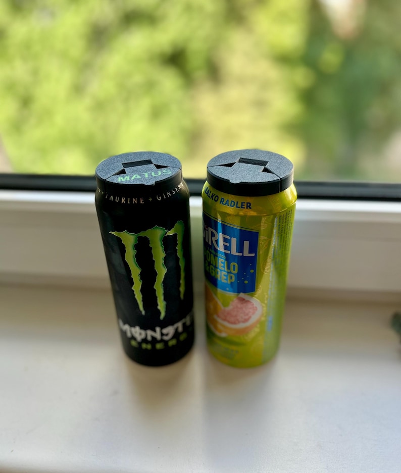 May include: Two aluminum beverage cans, one black with green claw marks and the word "MONSTER", the other yellow with the word "RELL". Both cans have black plastic lids. The background is a blurred view of green foliage.