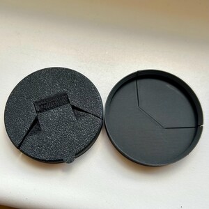 May include: Two-part black plastic object, likely a lid or cover, with a textured surface on one part and a smooth interior on the other. The parts are circular, with a segmented design on the interior. The textured part has a tab.