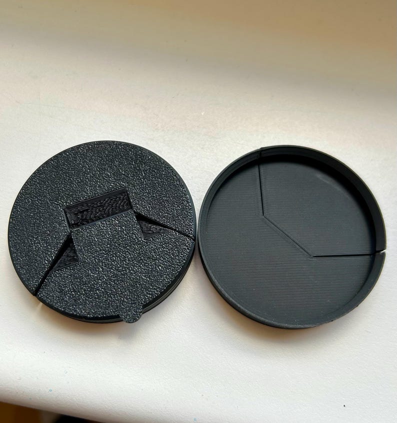 May include: Two-part black plastic object, likely a lid or cover, with a textured surface on one part and a smooth interior on the other. The parts are circular, with a segmented design on the interior. The textured part has a tab.