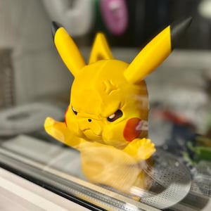 Peut inclure: Figurine Pikachu jaune avec des oreilles à pointes noires et des joues rouges. La figurine a une expression de colère et est posée avec un bras levé. La figurine est à l'intérieur d'une vitrine.