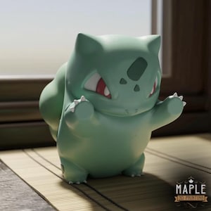 Puede incluir: Figura verde de Bulbasaur con ojos rojos y garras blancas, con los brazos levantados. La figura es una impresión 3D, como indica el logotipo de Maple 3D Printing. La figura está sobre una esterilla de bambú.