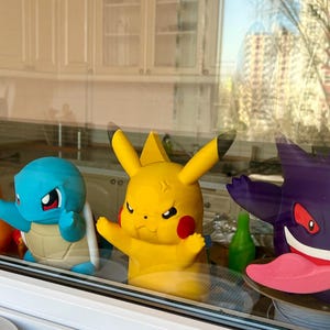 May include: A window display featuring four colorful 3D-printed Pokémon figures: Charmander, Squirtle, Pikachu, and Gengar. The figures are in vibrant orange, blue, yellow, and purple, with detailed features and expressions. The figures are displayed in front of a window.