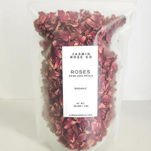 May include: A clear plastic bag filled with dried rose petals. The bag has a white label with the text "JASMIN ROSE CO" and "ROSES DRIED ROSE PETALS ORGANIC". The net weight is 28.356 grams / 1 oz. The website address is also printed on the label.