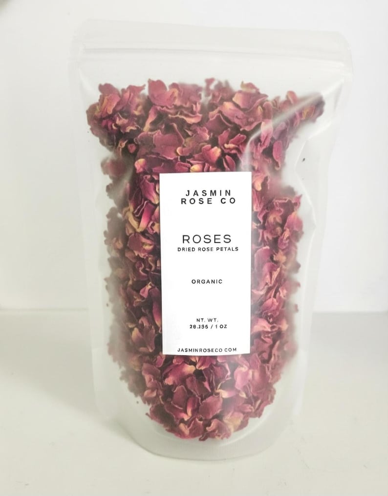 May include: A clear plastic bag filled with dried rose petals. The bag has a white label with the text "JASMIN ROSE CO" and "ROSES DRIED ROSE PETALS ORGANIC". The net weight is 28.356 grams / 1 oz. The website address is also printed on the label.