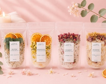 May include: Four clear pouches of luxurious bath soak, each with a different blend of ingredients. The pouches are labeled "Detox", "Energize", "Sunset", and "Soothe". Each pouch contains dried flowers and citrus slices. The background is a soft pink.