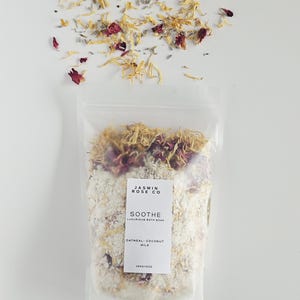 May include: A clear bag of bath soak with the text "JASMIN ROSE CO" and "SOOTHE LUXURIOUS BATH SOAK". The bag contains a white mixture with dried flowers and petals. The bag is topped with dried petals and flowers.