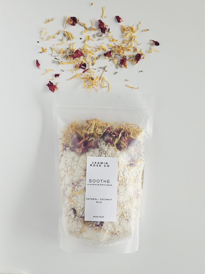 May include: A clear bag of bath soak with the text "JASMIN ROSE CO" and "SOOTHE LUXURIOUS BATH SOAK". The bag contains a white mixture with dried flowers and petals. The bag is topped with dried petals and flowers.