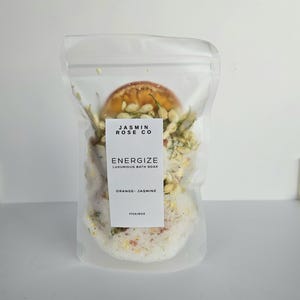May include: A clear, sealed bag containing bath soak. The bag features a white label with the text "JASMIN ROSE CO" and "ENERGIZE LUXURIOUS BATH SOAK ORANGE - JASMINE". The bag contains a mix of white granules, dried orange slices, and jasmine.