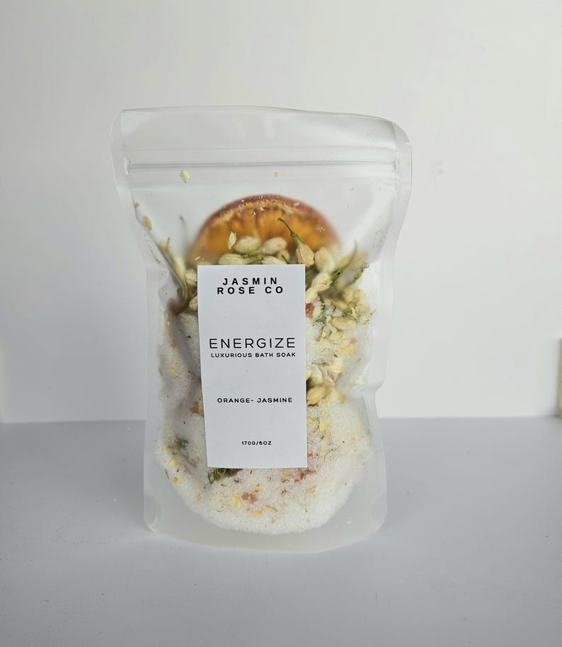 May include: A clear, sealed bag containing bath soak. The bag features a white label with the text "JASMIN ROSE CO" and "ENERGIZE LUXURIOUS BATH SOAK ORANGE - JASMINE". The bag contains a mix of white granules, dried orange slices, and jasmine.