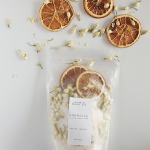 May include: A clear bag of bath soak with a label that reads "JASMIN ROSE CO" and "ENERGIZE LUXURIOUS BATH SOAK ORANGE - JASMINE". The bag contains white bath salts, dried orange slices, and jasmine buds. Scattered around are more orange slices and jasmine buds.