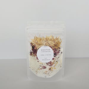 May include: A clear, resealable bag filled with oatmeal coconut milk bath soak. The bag contains dried flowers, rose petals, and white granules. A round label reads "SOOTHE Luxurious Bath Soak" and the website address.