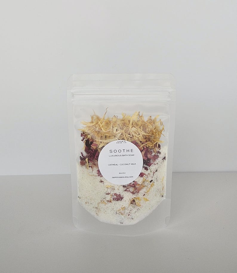 May include: A clear, resealable bag filled with oatmeal coconut milk bath soak. The bag contains dried flowers, rose petals, and white granules. A round label reads "SOOTHE Luxurious Bath Soak" and the website address.
