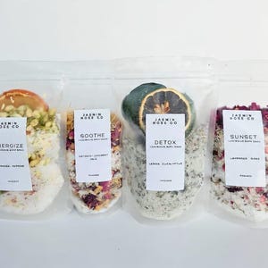 May include: Four clear plastic pouches filled with bath salts and botanicals. Each pouch has a label with the brand name and product name: Energize, Soothe, Detox, and Sunset. The pouches contain ingredients like orange, turmeric, oats, and rose petals.