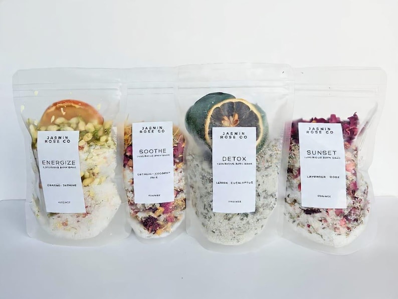 May include: Four clear plastic pouches filled with bath salts and botanicals. Each pouch has a label with the brand name and product name: Energize, Soothe, Detox, and Sunset. The pouches contain ingredients like orange, turmeric, oats, and rose petals.