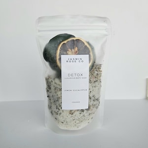 May include: A clear, resealable bag containing bath soak, a dried lemon slice, and a dark green object. The bag has a white label with the text "JASMIN ROSE CO" and "DETOX LUXURIOUS BATH SOAK LEMON - EUCALYPTUS". The bag is filled with a light-colored, granular substance.