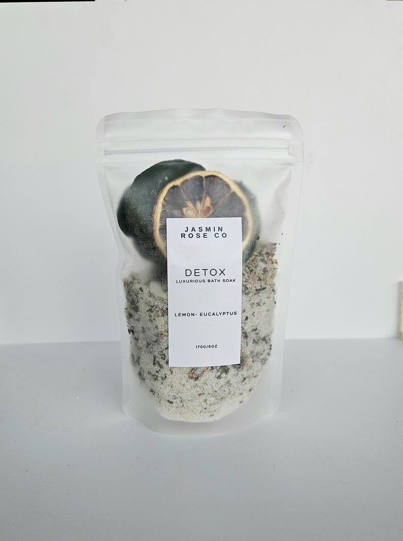 May include: A clear, resealable bag containing bath soak, a dried lemon slice, and a dark green object. The bag has a white label with the text "JASMIN ROSE CO" and "DETOX LUXURIOUS BATH SOAK LEMON - EUCALYPTUS". The bag is filled with a light-colored, granular substance.