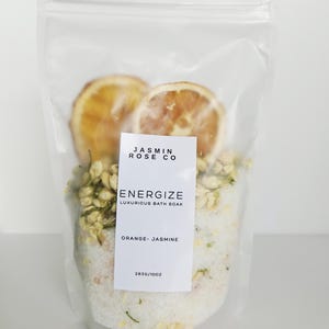 May include: A clear plastic bag filled with bath soak. The bag contains dried orange slices, jasmine flowers, and white bath salts. A white label reads "Energize Luxurious Bath Soak Orange-Jasmine".
