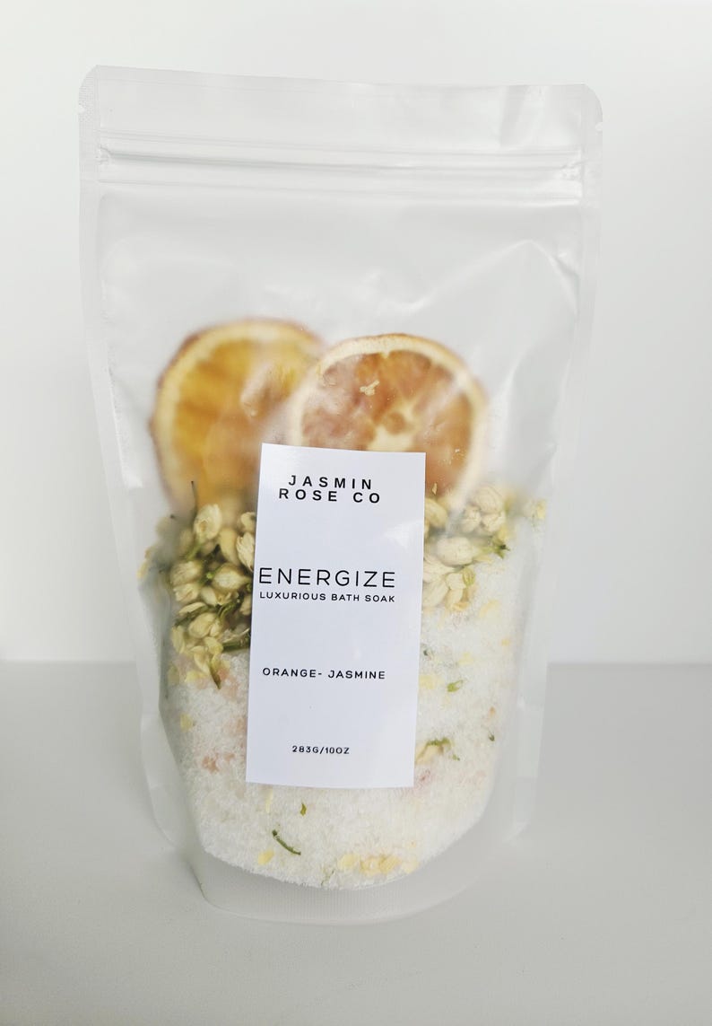 May include: A clear plastic bag filled with bath soak. The bag contains dried orange slices, jasmine flowers, and white bath salts. A white label reads "Energize Luxurious Bath Soak Orange-Jasmine".