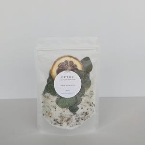 May include: A clear, resealable pouch filled with bath soak. The pouch contains white bath salts, dried lemon slices, and green eucalyptus leaves. A white circular label reads "DETOX LUXURIOUS BATH SOAK LEMON - EUCALYPTUS".