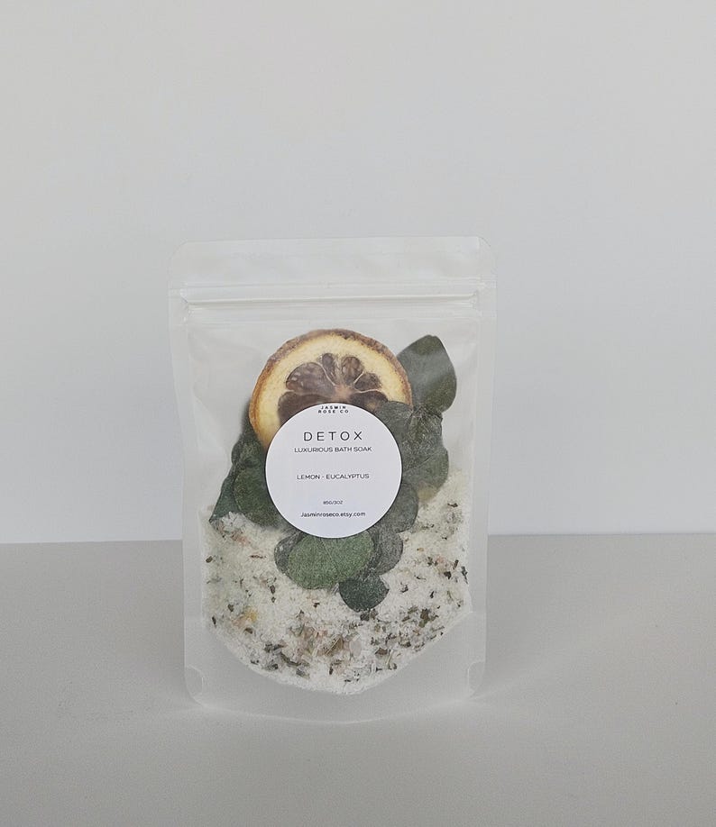 May include: A clear, resealable pouch filled with bath soak. The pouch contains white bath salts, dried lemon slices, and green eucalyptus leaves. A white circular label reads "DETOX LUXURIOUS BATH SOAK LEMON - EUCALYPTUS".