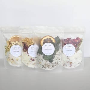 May include: Four clear pouches of luxurious bath soaks, each labeled with a different scent: Energize, Soo, Detox, and Sunset. Each pouch contains a blend of bath salts, dried botanicals, and a slice of dried fruit. The labels are white with black text.