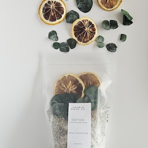 May include: A clear bag of bath soak with dried lemon slices and eucalyptus leaves. The bag has a label that reads "JASMIN ROSE CO DETOX LUXURIOUS BATH SOAK LEMON-EUCALYPTUS". The image also includes loose lemon slices and eucalyptus leaves.