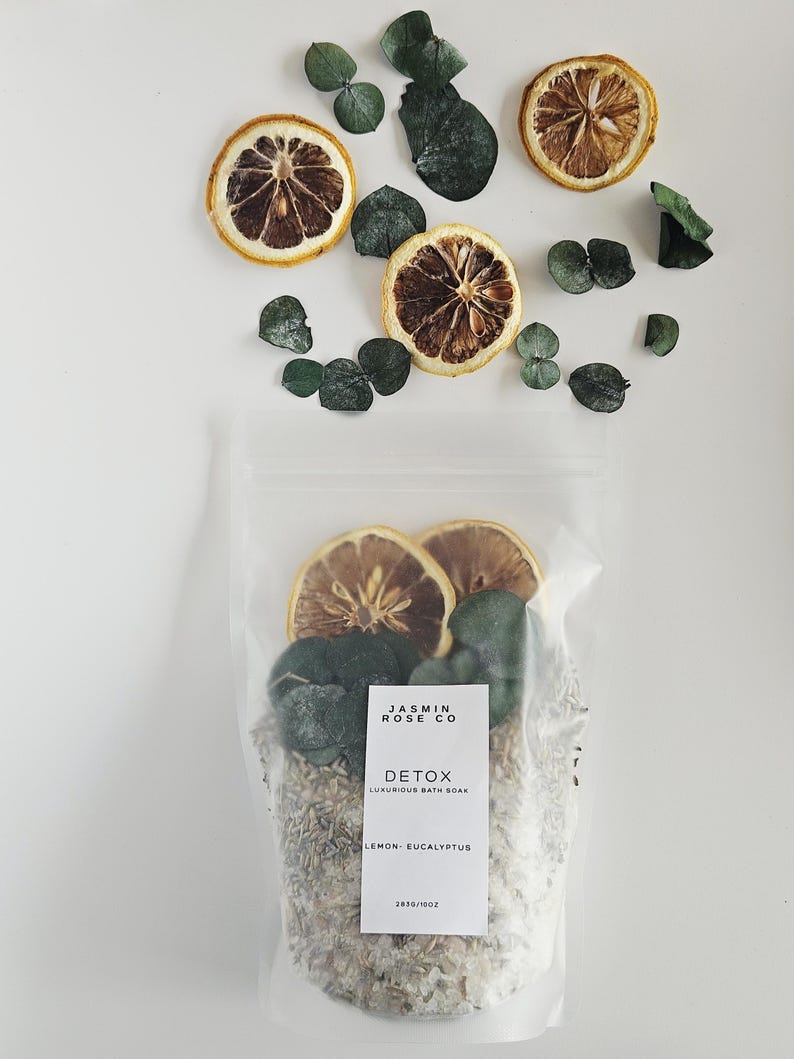 May include: A clear bag of bath soak with dried lemon slices and eucalyptus leaves. The bag has a label that reads "JASMIN ROSE CO DETOX LUXURIOUS BATH SOAK LEMON-EUCALYPTUS". The image also includes loose lemon slices and eucalyptus leaves.