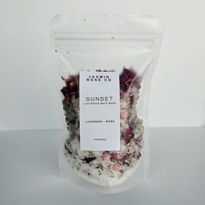 May include: A clear plastic bag filled with bath soak. The bag has a white label with the text "JASMIN ROSE CO" and "SUNSET LUXURIOUS BATH SOAK". The soak contains lavender, rose petals, and salt. The bag is labeled "170g/6oz".
