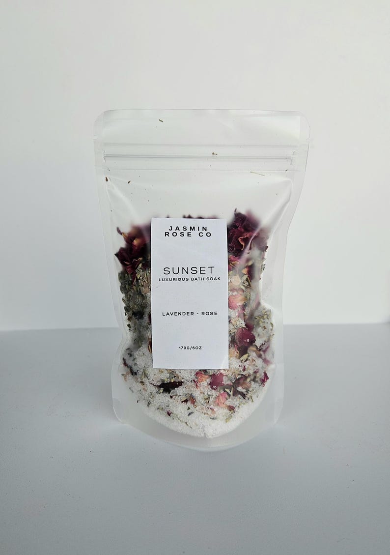 May include: A clear plastic bag filled with bath soak. The bag has a white label with the text "JASMIN ROSE CO" and "SUNSET LUXURIOUS BATH SOAK". The soak contains lavender, rose petals, and salt. The bag is labeled "170g/6oz".