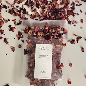 May include: A clear plastic bag filled with dried rose petals, with the text "JASMIN ROSE CO ROSES DRIED ROSE PETALS ORGANIC" on a white label. The net weight is 19.70 oz. Scattered rose petals surround the bag.