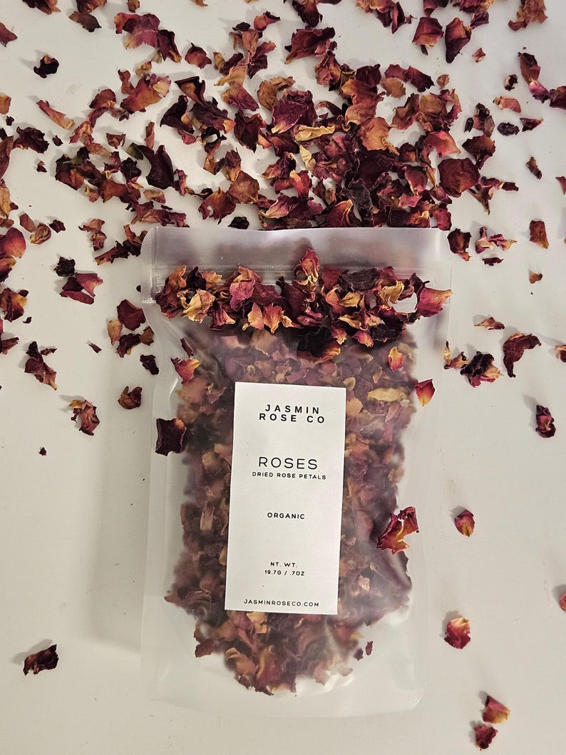 May include: A clear plastic bag filled with dried rose petals, with the text "JASMIN ROSE CO ROSES DRIED ROSE PETALS ORGANIC" on a white label. The net weight is 19.70 oz. Scattered rose petals surround the bag.