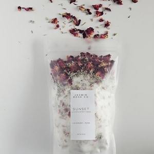 May include: A clear plastic bag filled with white bath salts, dried rose petals, and lavender. The bag has a white label that reads "Jasmin Rose Co" and "Sunset Luxurious Bath Soak Lavender - Rose". Scattered rose petals are above the bag.
