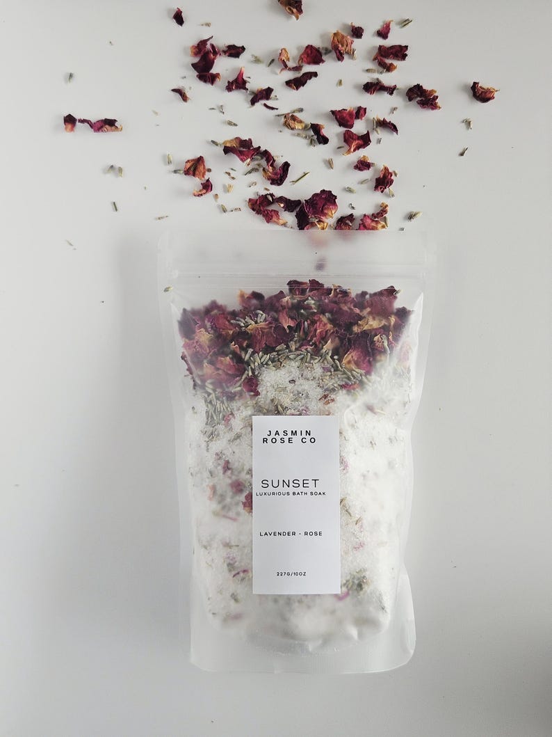 May include: A clear plastic bag filled with white bath salts, dried rose petals, and lavender. The bag has a white label that reads "Jasmin Rose Co" and "Sunset Luxurious Bath Soak Lavender - Rose". Scattered rose petals are above the bag.