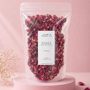 May include: A clear plastic bag filled with dried rose petals, labeled "ROSES DRIED ROSE PETALS ORGANIC" from Jasmin Rose Co. The bag sits on a pink pedestal, with loose petals scattered around. Net weight is 1 oz.