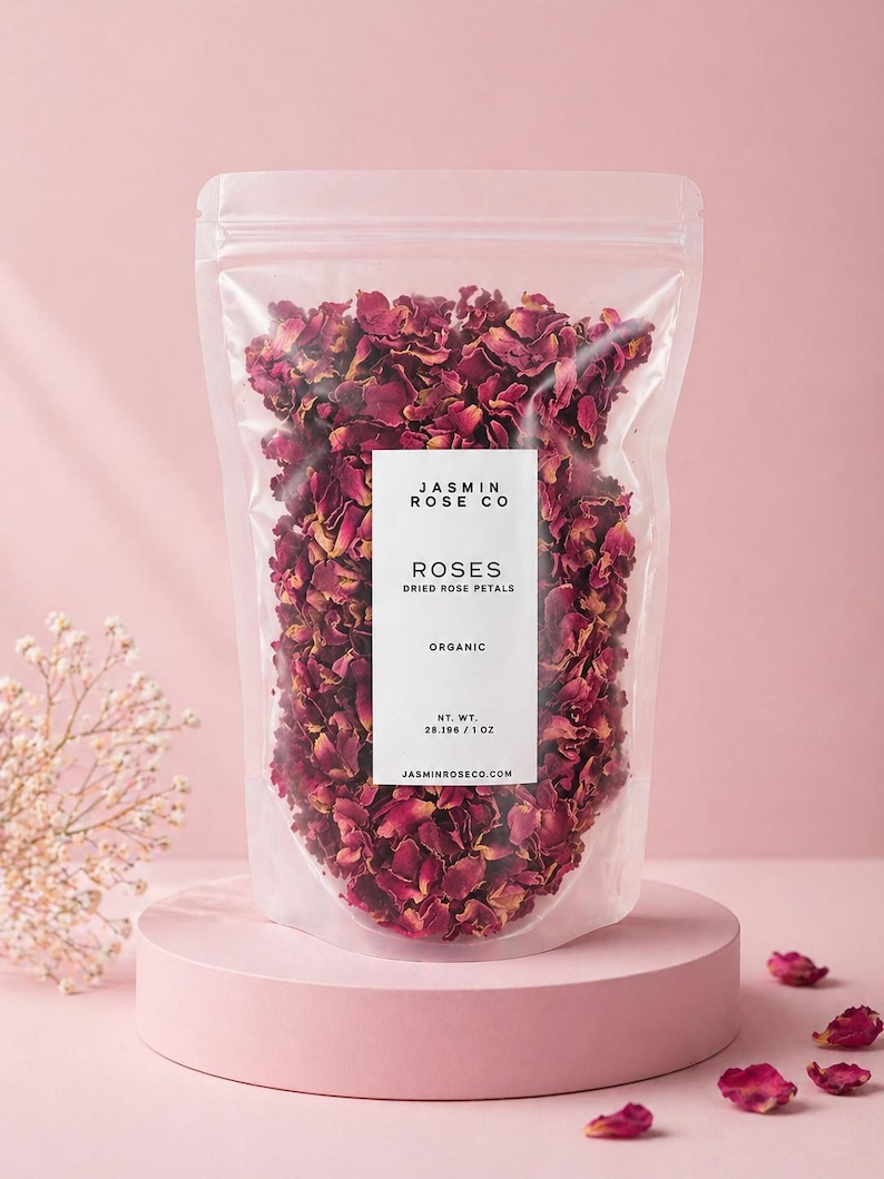 May include: A clear plastic bag filled with dried rose petals, labeled "ROSES DRIED ROSE PETALS ORGANIC" from Jasmin Rose Co. The bag sits on a pink pedestal, with loose petals scattered around. Net weight is 1 oz.