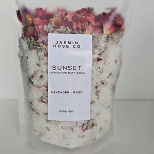 May include: A clear plastic bag filled with bath soak. The soak contains white granules, dried rose petals, and other botanicals. A white label reads "JASMIN ROSE CO" and "SUNSET LUXURIOUS BATH SOAK LAVENDER - ROSE". The bag contains 10 oz of product.