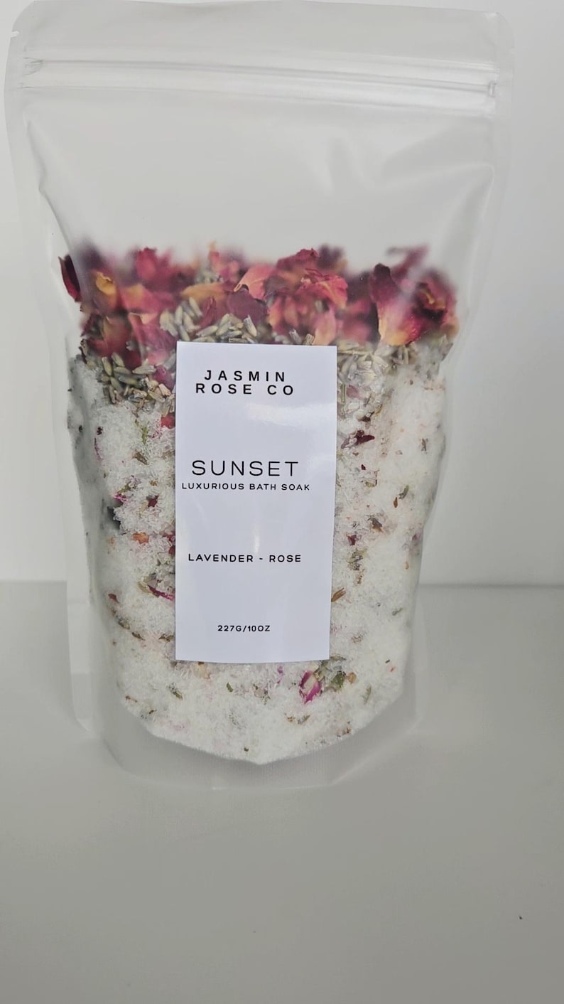 May include: A clear plastic bag filled with bath soak. The soak contains white granules, dried rose petals, and other botanicals. A white label reads "JASMIN ROSE CO" and "SUNSET LUXURIOUS BATH SOAK LAVENDER - ROSE". The bag contains 10 oz of product.