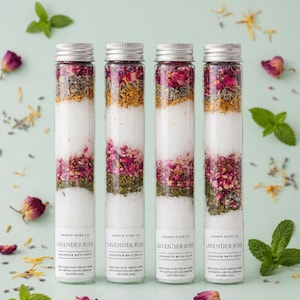 May include: Four glass tubes filled with layered bath salts. The layers include white salts, dried rose petals, herbs, and other botanicals. Each tube has a silver cap and a label that reads "Lavender Rose Luxurious Bath Salts."