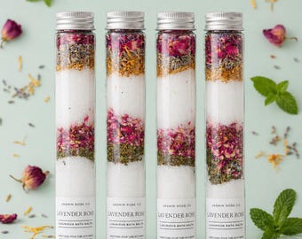 May include: Four glass tubes filled with layered bath salts. The layers include white salts, dried rose petals, herbs, and other botanicals. Each tube has a silver cap and a label that reads "Lavender Rose Luxurious Bath Salts."
