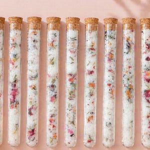 May include: Nine clear glass tubes filled with white granules and colorful dried flowers and herbs, each sealed with a cork stopper. The tubes are arranged in a row against a soft pink background, suggesting a spa or beauty product.