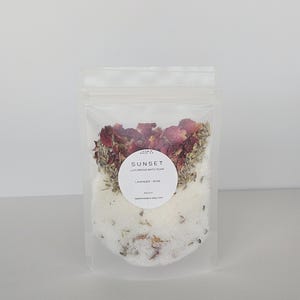 May include: A clear, resealable bag filled with a luxurious bath soak. The product is labeled "Sunset Luxurious Bath Soak Lavender - Rose." The soak contains dried rose petals, lavender buds, and white bath salts. The bag is displayed against a plain white background.